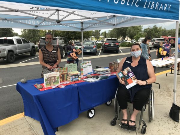Library staff, one standing and one seated in a wheelchair, work at the library's table on the sidewalk at back to school night during the COVID-19 pandemic.
