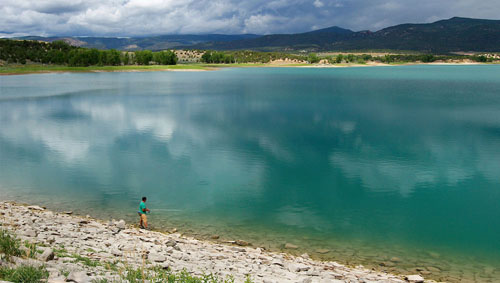 A large turquoise lake in the foreground with trees, mountains, and a cloudy sky. A person is fishing on the shore of the lake.