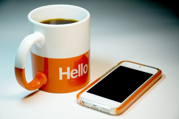 An iphone sits on a white table next to a n orange and white coffee mug with the word "Hello" inscribed on it.